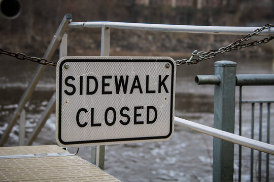 Close-up of a Sidewalk Closed sign on a metal gate, with rushing river water and stairs in a blurred background. Ideal for themes of safety, construction, detour, or infrastructure maintenance.