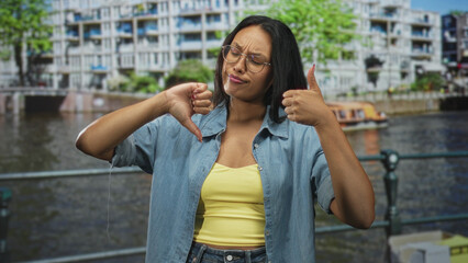 Fototapeta premium Woman showing thumbs up and thumbs down with visible hands and smile at canal railing in front of apartment building; ambivalence.