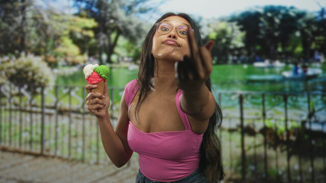 Woman in pink top and glasses holding a multicolored ice cream cone flips middle finger by a lakeside fence in street; sassy defiance.