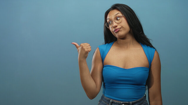 Young woman points thumb over shoulder in studio wearing glasses and blue sleeveless top and denim jeans; indifference sarcasm.