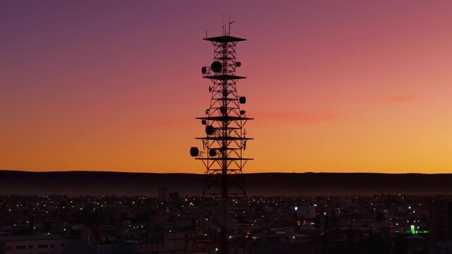 Durango, Mexico cityscape at dusk with tall communication tower silhouetted against gradient orange pink and purple sunset sky. City lights sparkle below distant mountains on horizon.