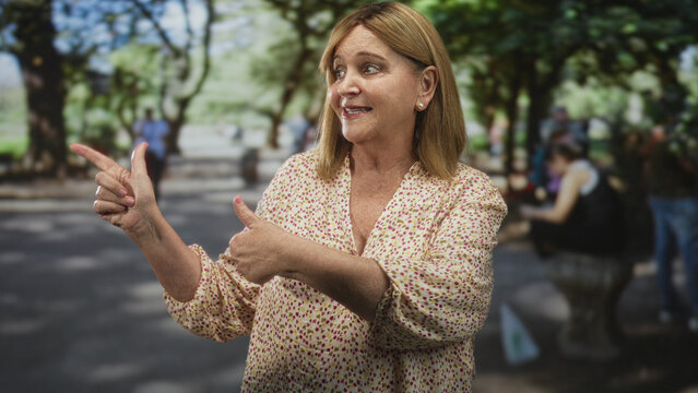 Middle age woman points finger and gestures thumb toward self on a street with trees and benches; cheerful friendly conversation.