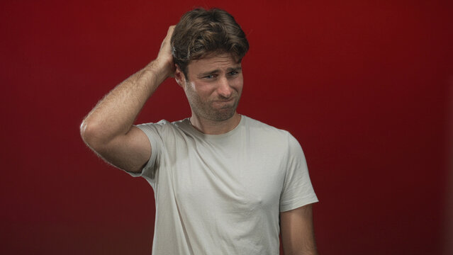 Young hispanic man with hand on back of head and furrowed brow wearing white tshirt in a red studio showing a puzzled scratching gesture and slouched posture; confusion.