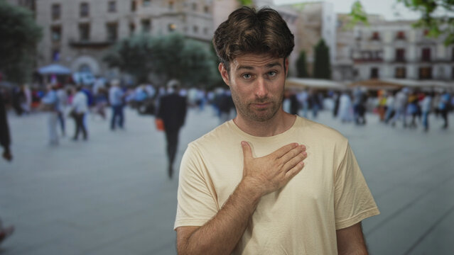 Man with hand on chest on crowded street in front of plaza buildings, gesture of apology and modesty while facing camera; sincere regret.
