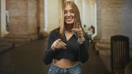Obraz premium Redhead woman pointing finger and showing bare midriff beneath a black cropped top in a stone building archway with passerby blur; youthful confidence.