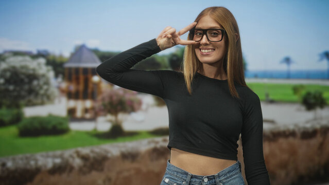 Woman redhead wearing glasses giving peace sign to eye and showing midriff in a black crop top and jeans on a street by a seaside pavilion; playful joy.