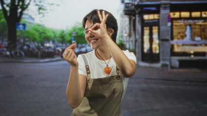 Fototapeta premium Woman holds ring in hand and makes ok sign with fingers near eye in street storefront setting; joyful curiosity.