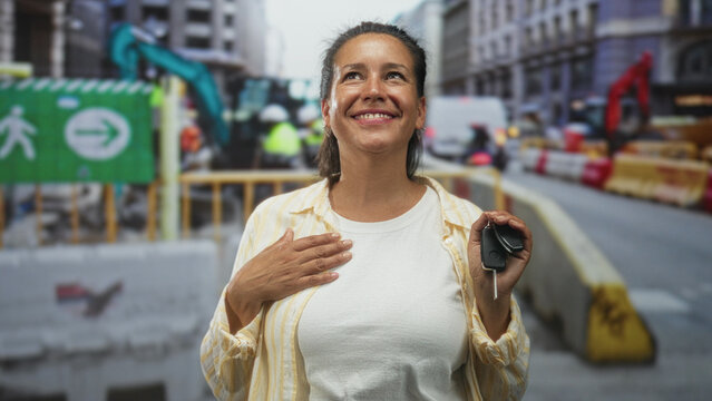 Woman holding car keys, hand to chest smiling on a busy urban street with construction barriers, bus, van and pedestrian sign, wearing white tee and striped shirt; joy.