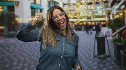 Fototapeta premium Woman gives thumbs up with visible hands and smiling face on a busy street with festive lights and storefronts; cheerful optimism.