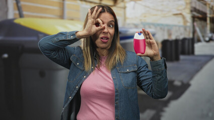 Fototapeta premium Middle aged hispanic woman holds pink toothpaste bottle near face, makes ok sign to eye and smiles in airport baggage claim carousel area; playful confidence.