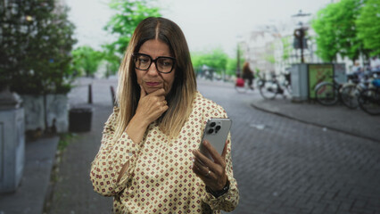 Fototapeta premium Middle aged woman holding smartphone and touching chin, looking puzzled amid bicycles on a cobblestone street; doubt.