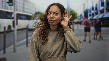 Fototapeta premium Young woman cupping her ear with hand on a busy city street beside buses and shops listening for a sound or voice; curiosity.