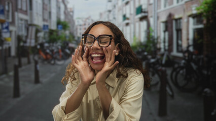 Fototapeta premium Woman with hands cupped around mouth and glasses on street lined with bicycles and narrow buildings; joyful playfulness.