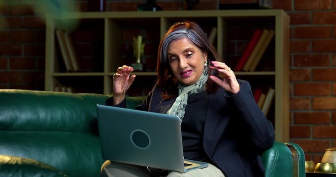 Indian asian senior businesswoman in corporate attire working on laptop at workstation in modern cozy office space experienced female executive with white hair sitting at professional desk area