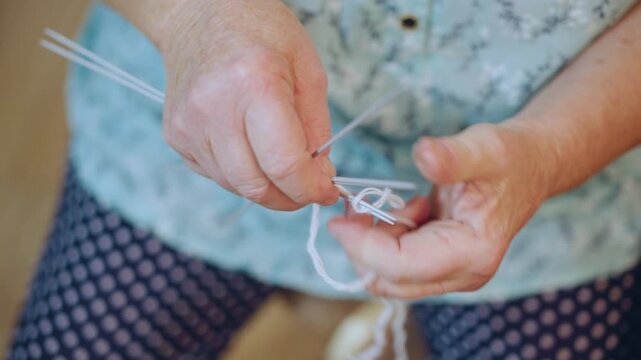 Closeup white grandma hands knitting needles, veteran seamstress casting stitches with soft white yarn on polkadot lap, floral blouse visible, warm natural light, cozy nostalgic atmosphere