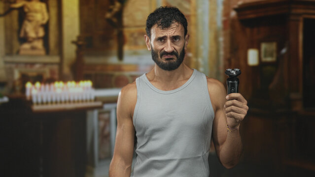 Man holding electric shaver with beard visible and grimace beside votive candles at a church building; unease.
