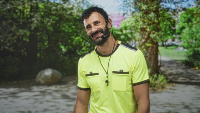 Man referee smiling with whistle and beard, wearing yellow referee jersey and headset on a forest path with trees and a rock; fairness friendly.