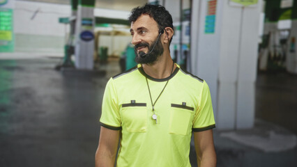 Man in neon yellow shirt with whistle on lanyard and headset smiles and tilts head by gas station forecourt pumps, beard visible  friendly service. © Krakenimages.com