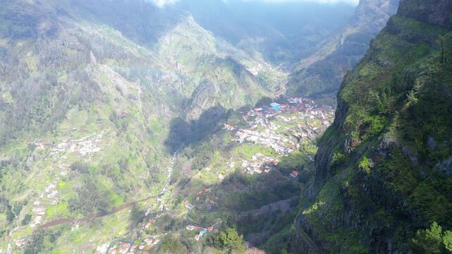 Aerial view of Curral das Freiras, Madeira, nestled in a deep valley surrounded by verdant mountains. A modern tourist complex is visible.
