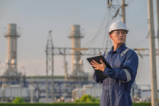 Asian male electrical engineer holding digital tablet standing at power plant station. Professional energy industry technician working on infrastructure maintenance and renewable resource project.