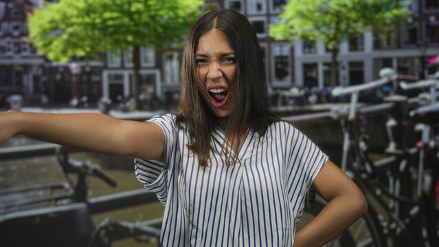 Woman pointing arm outward and sticking out tongue on street near canal railing and bicycles; playful defiance.