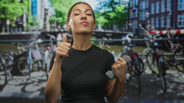 Young brunette woman holding a small dumbbell in one hand and giving thumbs up with the other on amsterdam street; confidence.