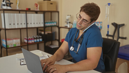 Fototapeta premium Man doctor with stethoscope wearing blue scrubs and glasses holds phone to ear and types on laptop in building; focused care.