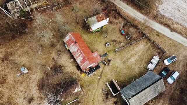 Aerial View Rural House Yard Rusted Red Roof Scattered Vehicles Small Outbuilding Dirt Road Leafless Trees