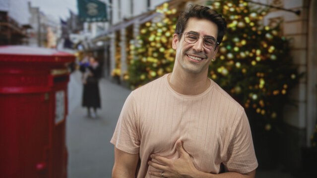 Man with hand on chest smiling and looking up, wearing glasses and a ribbed t-shirt, standing by a red postbox on a christmas decorated city street; holiday joy.