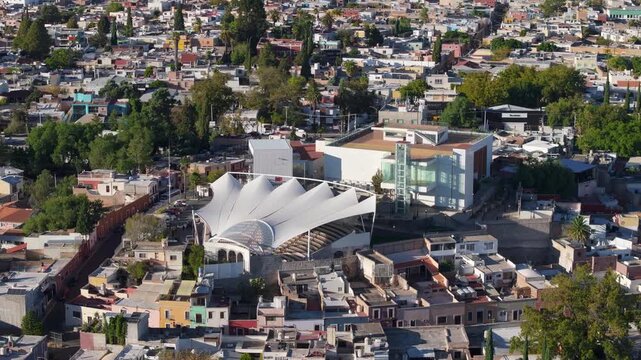 Aerial view of a modern open-air amphitheater featuring distinctive white tensile fabric roof structure surrounded by colorful residential neighborhoods in Durango Mexico during golden hour sunlight.
