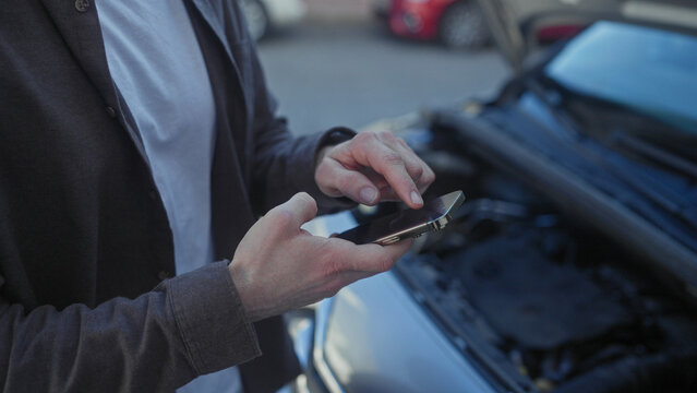 Man using smartphone with hands near open car hood and engine compartment on street; concern assistance.