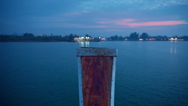 Weathered rustic dock post with rusted wooden panel and metal frame stands in foreground as calm blue waters stretch toward distant shore lights glowing under pink and blue twilight sky at dusk.