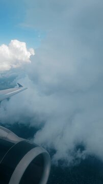 Vertical Screen: Airplane wing and jet engine visible through passenger window as aircraft flies through dense cloud formations with patches of blue sky and white cumulus clouds above.