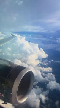 Vertical Screen: Airplane jet engine and wing soar high above dramatic white cumulus clouds against a vibrant blue sky, with sunlight streaming through the cabin window creating soft lens flares.