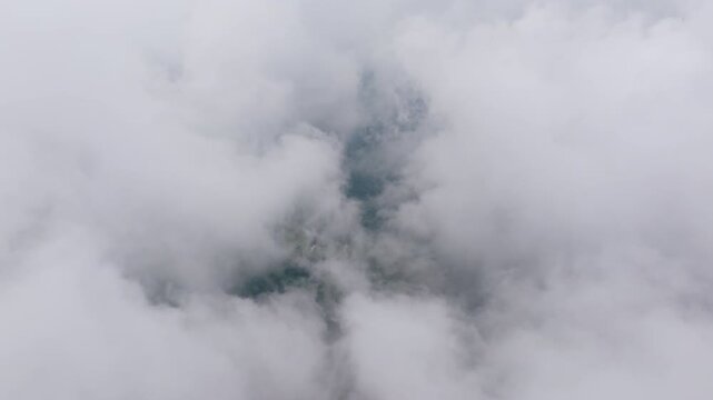 Aerial drone footage captures thick white clouds and mist slowly drifting and swirling above a lush green forested mountainside, with glimpses of terrain visible through breaks in the fog layer.
