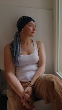 Woman undergoing chemotherapy sitting by window at home, headscarf cancer patient looking outside in reflection, recovery journey, resilience, solitude, hope and emotional healing