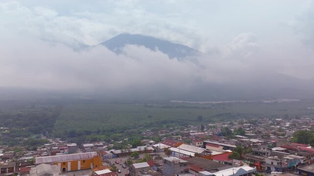 Drone footage captures a sprawling Guatemalan town with colorful rooftops nestled against a dramatic volcanic mountain shrouded in clouds and mist. Green agricultural fields stretch toward foggy.