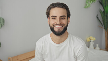 Obraz premium Man on bed smiling, face and beard visible, wearing white tshirt and gold chain, facing camera in studio; relaxed contentment.