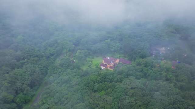 Drone footage reveals secluded villa with terracotta roof nestled in dense tropical rainforest as thick fog rolls across lush green mountain landscape creating mystical atmosphere.