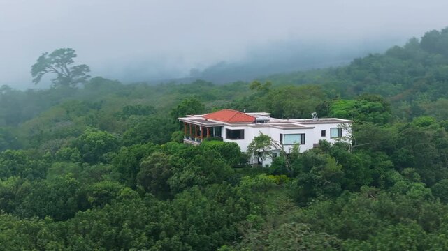 Aerial view of modern white mansion with terracotta roof surrounded by dense tropical rainforest. Fog rolls over lush green mountains creating serene atmosphere in remote location.