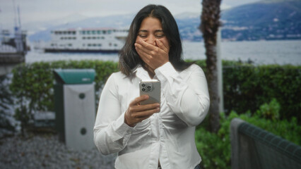 Fototapeta premium Woman holding smartphone and covering mouth after reading a message at a lakeside promenade with ferry and palm trees behind her; surprise connection.