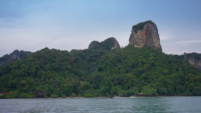 Traditional Thai longtail boat cruises across emerald waters as dramatic limestone karst cliffs covered in lush green jungle rise majestically against an overcast sky in Krabi, Thailand.