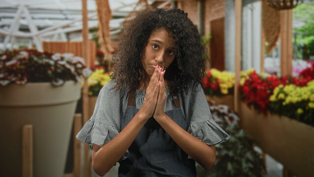 Woman in apron presses hands together in pleading prayer gesture while pouting lips among potted flowers and greenhouse plants inside a building; apologetic plea.