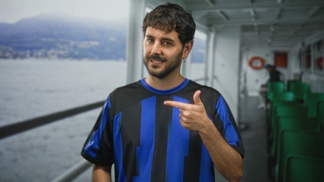 Man points finger at blue football shirt on a ferry deck cabin inside a building, smiling and showing hand gesture; confidence team spirit.