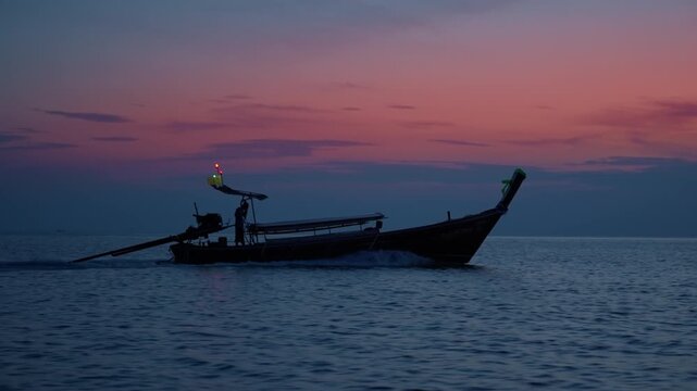 Silhouette of traditional Thai longtail boat with glowing navigation lights gliding across dark rippling ocean waters under dramatic pink and purple gradient sunset sky during peaceful dusk hours.