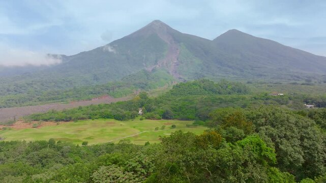 Aerial view of active Fuego Volcano in Guatemala rising above dense tropical rainforest. Lush green vegetation covers the valley with twin volcanic peaks against cloudy sky and misty atmosphere.