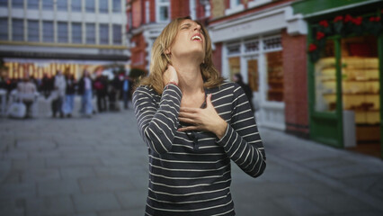 Fototapeta premium Woman clutching chest and pressing neck with hand in a busy urban shopping street by storefronts and crowds; pain urgency concern.