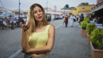 Fototapeta premium Woman in yellow tank top gesturing hand toward camera with exposed midriff on a busy street market; playful confidence.