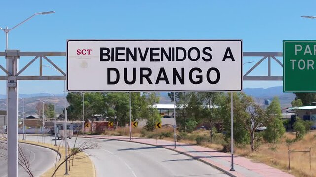 Welcome to Durango Mexico road sign on highway entrance with distant mountains and clear blue sky. Street lamps line the empty road with green trees and dry grass visible in the sunny afternoon.