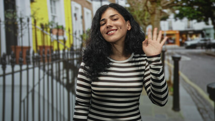 Young arab woman with curly dark hair waving right hand on city street near iron railing and tree...